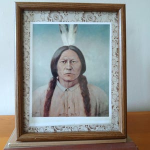 May include: Framed portrait of Sitting Bull, featuring a detailed depiction of the Native American leader. The portrait is set within a wooden frame with a floral patterned border. The image rests on a vintage book titled "BEAUTYWAY A NAVAHO CEREMONIAL".