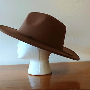 May include: A brown fedora hat with a wide brim, displayed on a white mannequin head. The hat has a classic shape with a slightly indented crown and a ribbon band around the base of the crown. The hat is made of a felt-like material.