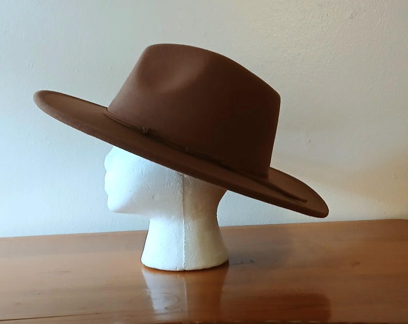 May include: A brown fedora hat with a wide brim, displayed on a white mannequin head. The hat has a classic shape with a slightly indented crown and a ribbon band around the base of the crown. The hat is made of a felt-like material.