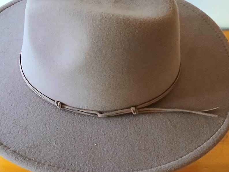 May include: A light brown fedora-style hat with a wide brim and a matching band around the crown. The hat has a soft, felt-like texture and a classic, timeless design. The hat is suitable for various occasions.