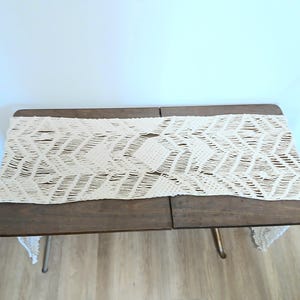 May include: A wooden table with a cream-colored macrame table runner. The table has a dark brown finish and metal legs. The macrame runner features a geometric pattern.