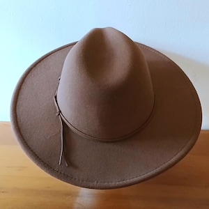 May include: A brown fedora-style hat with a wide brim and a leather band around the crown. The hat has a teardrop crown and a slightly curved brim. The hat is made of a soft material and is suitable for various occasions.