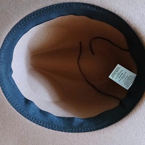 May include: A tan-colored fedora hat with a dark blue brim and a brown interior. The hat has a classic design, with a structured crown and a slightly curved brim. A tag is visible inside the hat.