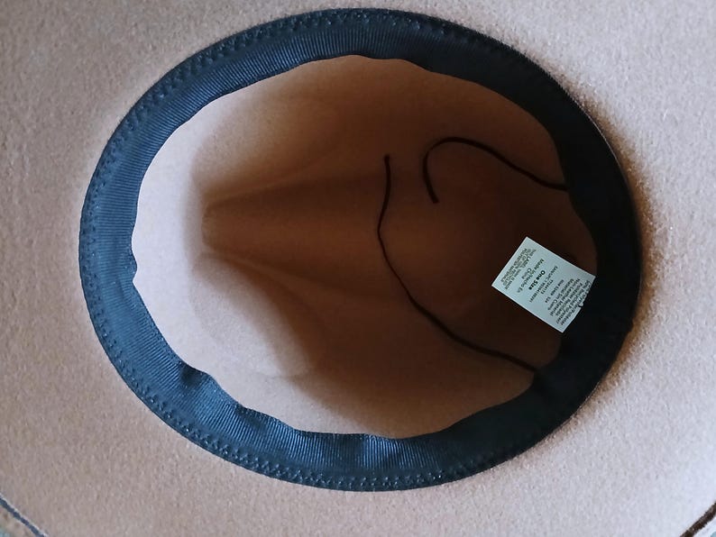 May include: A tan-colored fedora hat with a dark blue brim and a brown interior. The hat has a classic design, with a structured crown and a slightly curved brim. A tag is visible inside the hat.