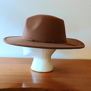 May include: A brown fedora-style hat with a wide brim and a decorative band. The hat is displayed on a white mannequin head, set against a light background. The hat's brim measures approximately 3 inches.