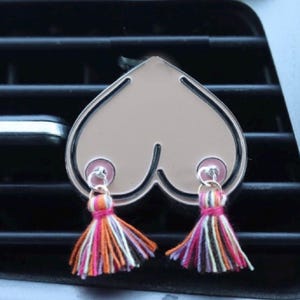 May include: A heart-shaped car air freshener or decorative accessory with a metallic silver finish. The design features two circular pink accents at the bottom, each attached to a small, multi-coloured fabric tassel with orange, pink, and black threads.