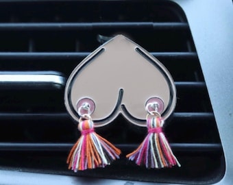 Pode incluir: Um ambientador de carro ou acessório decorativo em forma de coração com um acabamento prateado metálico. O design apresenta dois detalhes circulares cor-de-rosa na parte inferior, cada um preso a uma pequena borla de tecido multicolorida com fios laranja, rosa e pretos.