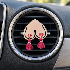May include: A car air freshener vent clip featuring a light beige heart-shaped enamel piece with black outlines. Two small pink heart-shaped charms are attached to the bottom, each featuring a dangling bright pink fabric tassel with silver accents.
