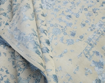 Könnte beinhalten: Eine aufgerollte, cremefarbene Teppich mit einem detaillierten Blumenmuster in Blautönen. Die Ränder des Teppichs sind mit einer blauen Franse versehen. Das Design weist einen klassischen, verzierten Stil auf, der sich für die Inneneinrichtung eignet.