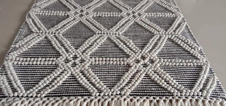 May include: A white and black geometric rug with a diamond pattern. The rug has a fringe border and is made of a textured material.Chunky weave wool rug, natural wool rug, mix weave wool rug, boho home decor rug, indoor rug,