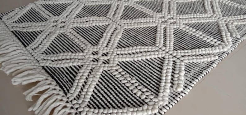 May include: A black and white woven rug with a geometric pattern and fringe edges. The rug has a textured, raised surface.Chunky weave wool rug, natural wool rug, mix weave wool rug, boho home decor rug, indoor rug,