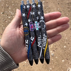 May include: Four black glitter pens with different colored stripes and designs. The pens have the names "Mark", "Miller", and "Miller" written on them.
