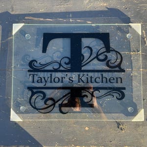 Puede incluir: Una tabla de cortar de vidrio con una calcomanía de vinilo negro de una letra "T" grande con adornos decorativos y el texto "Taylor's Kitchen".