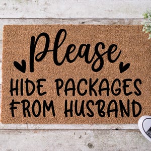 May include: A rectangular doormat made of natural coir fiber. The mat features the words "Please HIDE PACKAGES FROM HUSBAND" in black script. Two black hearts flank the word "Please". Black and white sneakers are visible in the bottom right corner.