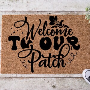 Puede incluir: Una felpudo rectangular de fibra de coco con las palabras "Welcome to Our Patch" en escritura negra. La alfombra tiene un color marrón natural y está decorada con una calabaza y estrellas. Zapatillas negras y blancas visibles en la esquina inferior derecha.