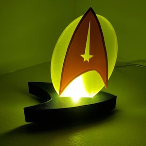 Może przedstawiać: Nocna lampka w kształcie żółto-czerwonego emblematu Star Trek z czarną podstawą. Światło jest włączone i oświetla otaczającą przestrzeń.