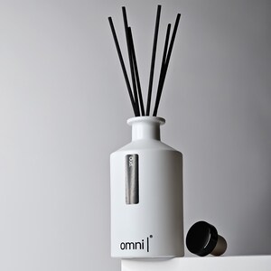 Pode incluir: Difusor de óleos essenciais em cerâmica branca com palhetas pretas. O difusor tem formato cilíndrico com gargalo estreito e um rótulo preto que diz "Oud". A palavra "omni" está impressa na frente. Uma rolha preta fica ao lado do difusor.