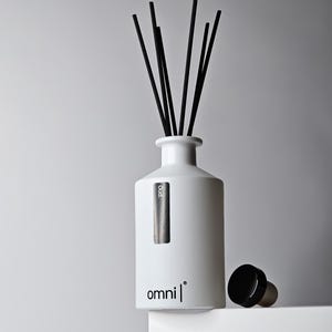 May include: A white ceramic reed diffuser bottle with a matte finish, featuring the text "omni" and a vertical label reading "Oud." Six thin black reeds extend from the neck. A separate black stopper with a cork base sits beside the bottle.