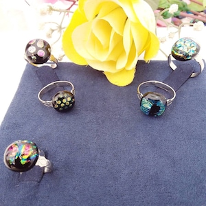 May include: Four rings with circular black glass cabochons featuring iridescent, multi-colored flecks and patterns. Each ring has a thin, silver-toned metal band. The glass surfaces display reflective specks of pink, blue, green, and gold colors.