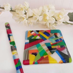 May include: A set of two colorful glass coasters with an abstract design. The coasters are made of fused glass and feature a variety of colors, including red, yellow, green, blue, and white.