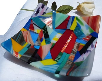 May include: A square glass dish with a mosaic design. The dish is made of many small pieces of glass in various colors, including red, blue, yellow, green, and white. The glass pieces are arranged in a geometric pattern.
