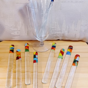 May include: A collection of clear glass stirring sticks with multicolored mosaic-style tips. Five sticks are standing upright inside a clear glass goblet, while eight additional sticks are arranged horizontally in front of the glass.