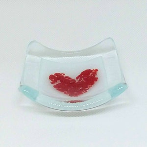 May include: A small, clear glass dish with a curved shape. The dish has a red heart design in the center.