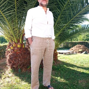 Puede incluir: Una persona con una camisa blanca y pantalones de lino beige con cintura con cord&oacute;n. Lleva sandalias azules. El fondo incluye una palmera y hierba verde.