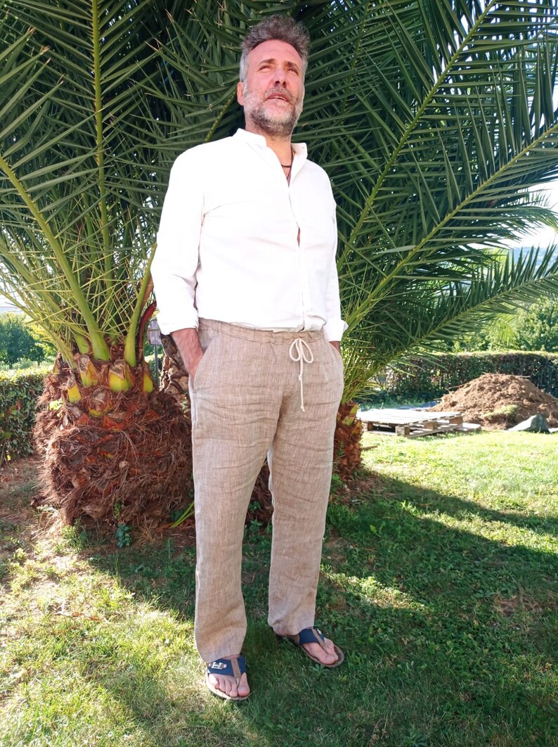 Puede incluir: Una persona con una camisa blanca y pantalones de lino beige con cintura con cord&oacute;n. Lleva sandalias azules. El fondo incluye una palmera y hierba verde.