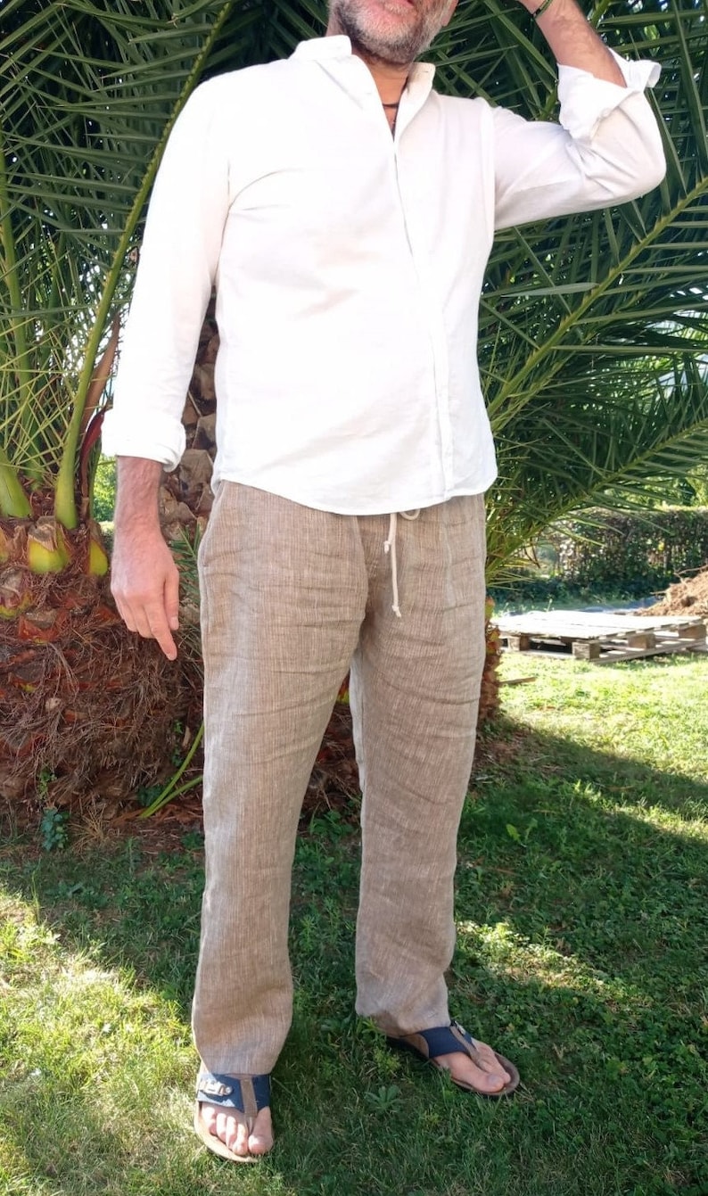 Puede incluir: Una persona con una camisa blanca y pantalones de lino color beige. Los pantalones tienen cintura con cord&oacute;n. La persona tambi&eacute;n lleva sandalias. El atuendo es adecuado para el clima c&aacute;lido.