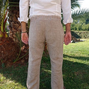 Puede incluir: Pantalones de lino color beige con un corte holgado, combinados con una camisa blanca de manga larga. Los pantalones tienen un dise&ntilde;o de pierna recta. La persona est&aacute; de pie sobre hierba, con una palmera al fondo.