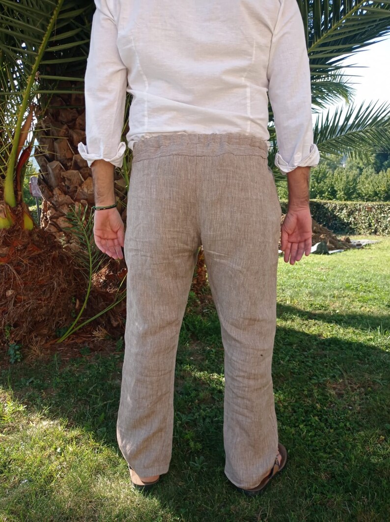 Puede incluir: Pantalones de lino color beige con un corte holgado, combinados con una camisa blanca de manga larga. Los pantalones tienen un dise&ntilde;o de pierna recta. La persona est&aacute; de pie sobre hierba, con una palmera al fondo.