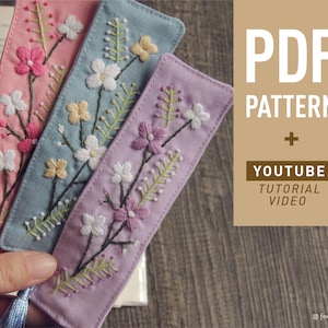May include: Three embroidered bookmarks with floral designs in pink, blue, and purple. The bookmarks have tassels and are displayed on a book. The image includes the text "PDF PATTERN + YOUTUBE TUTORIAL VIDEO".