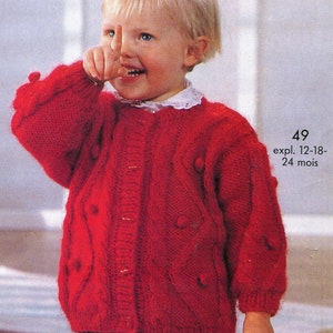 May include: A red knitted cardigan with a cable knit pattern and button closure. The cardigan has a white lace collar and is decorated with small pom-poms. The cardigan is for a child aged 12-18 months or 24 months. The text on the image reads "49 expl. 12-18-24 mois".