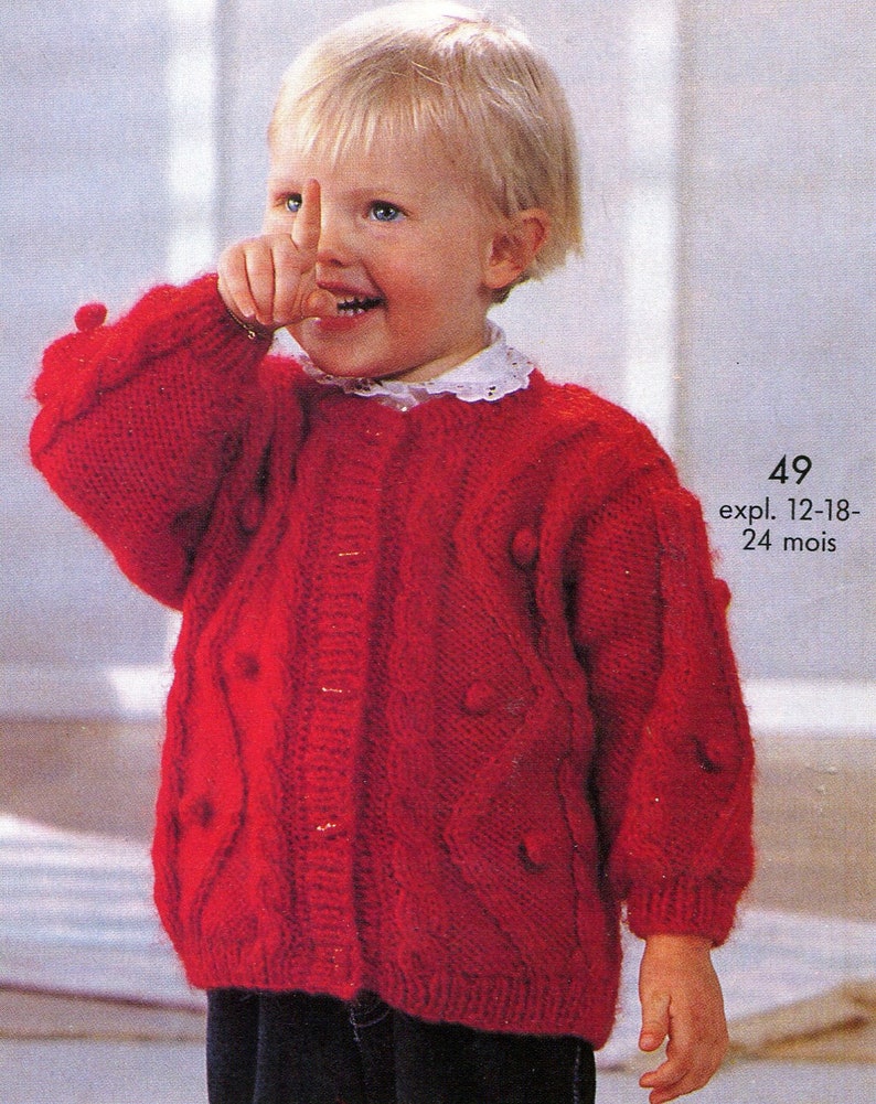 May include: A red knitted cardigan with a cable knit pattern and button closure. The cardigan has a white lace collar and is decorated with small pom-poms. The cardigan is for a child aged 12-18 months or 24 months. The text on the image reads "49 expl. 12-18-24 mois".