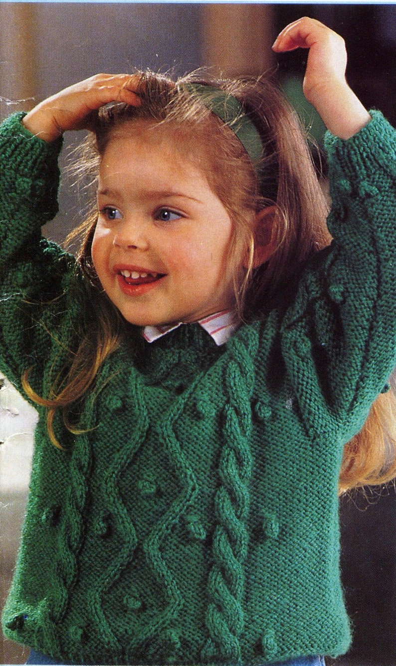 May include: A young girl wearing a green knitted sweater with a cable knit pattern and a green headband. The sweater has a textured surface and is decorated with small, round, white buttons.