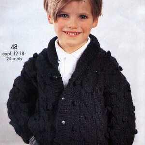May include: A black knitted cardigan with a cable knit pattern and button closure. The cardigan has a collar and is worn by a young boy with blonde hair. The text "48 expl. 12-18-24 mois" is visible on the image.