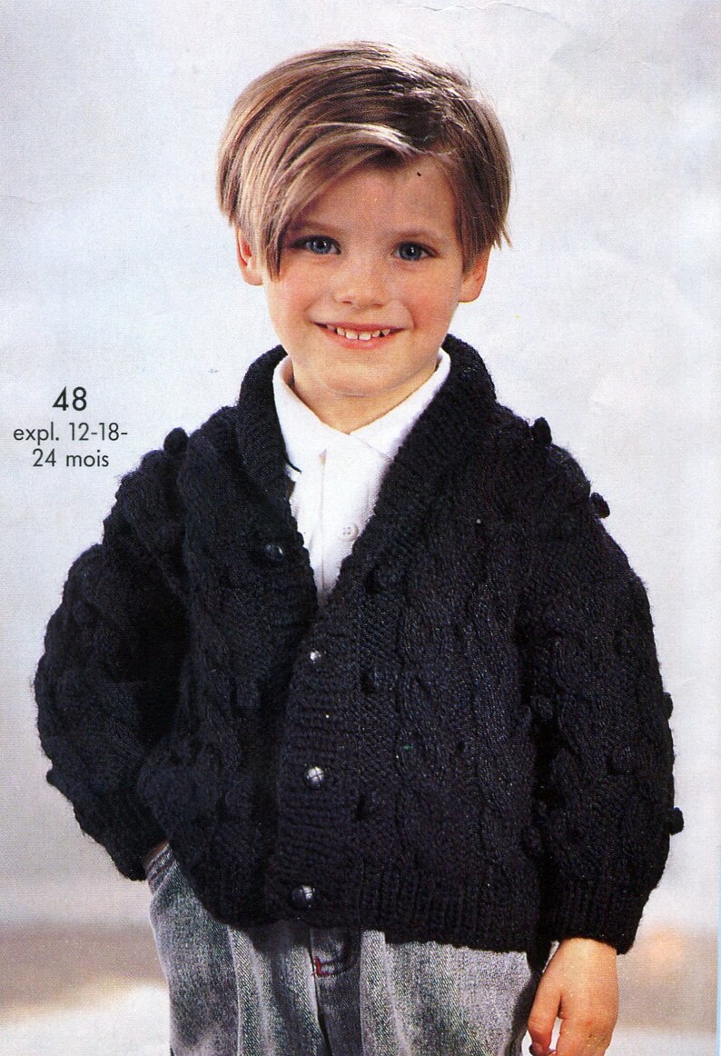 May include: A black knitted cardigan with a cable knit pattern and button closure. The cardigan has a collar and is worn by a young boy with blonde hair. The text "48 expl. 12-18-24 mois" is visible on the image.