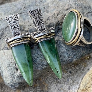Op de afbeelding: Een set groene jade sieraden met twee hangers en een ring. De hangers zijn zilver met gouden accenten en de ring is zilver met een gouden band. De jade is diep groen van kleur met zwarte vlekken.