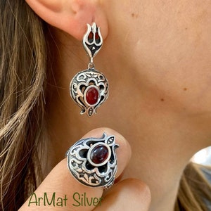 Può includere: Un anello e orecchini d'argento con una pietra rossa a forma di melograno. L'anello e gli orecchini presentano intricati disegni floreali.