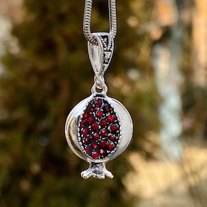 May include: A silver chain necklace with a pomegranate pendant. The pendant is made of silver and has red gemstones that resemble pomegranate seeds.