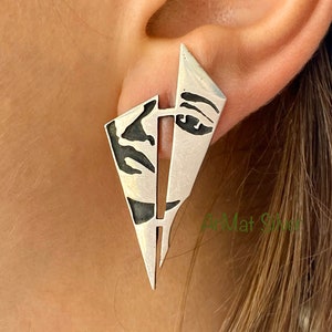 May include: Silver earrings in the shape of a triangle with a face design. The earrings are a geometric design with a black and white face silhouette.