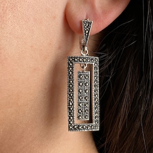 May include: Silver dangle earrings with a rectangular design featuring a smaller rectangle inside. The earrings are made with marcasite stones.
