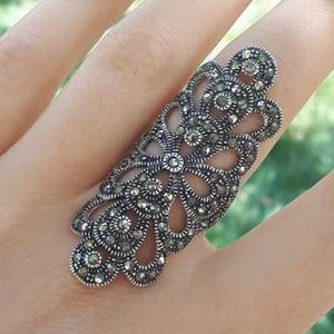 Op de afbeelding: Een zilveren ring met een delicaat, ingewikkeld ontwerp met een cluster van kleine, zwarte edelstenen. De ring wordt gedragen op de middelvinger van een hand.