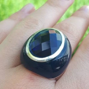 Puede incluir: Un anillo grande, azul oscuro con una piedra facetada de forma ovalada. El anillo tiene una banda plateada alrededor de la piedra y una base azul oscuro. El anillo está en la mano de una persona, con un fondo verde.