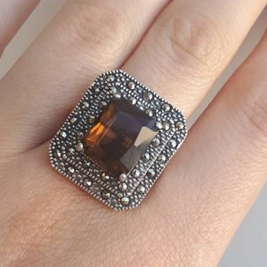 Puede incluir: Un anillo de plata con una piedra preciosa marrón de forma cuadrada rodeada de pequeñas piedras plateadas.