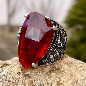 Peut inclure: Une bague en argent avec une grande pierre précieuse rouge facetée. La bague présente un design filigrané détaillé. ArMat Silver est imprimé sur le rocher sous la bague.