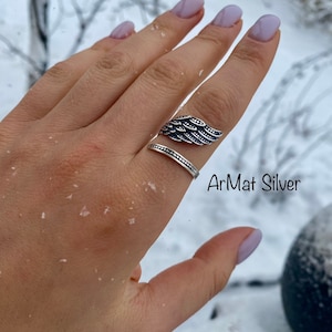 May include: A silver ring with a detailed wing design. The ring is worn on a finger with light pink nail polish. The text "ArMat Silver" is visible in the image.