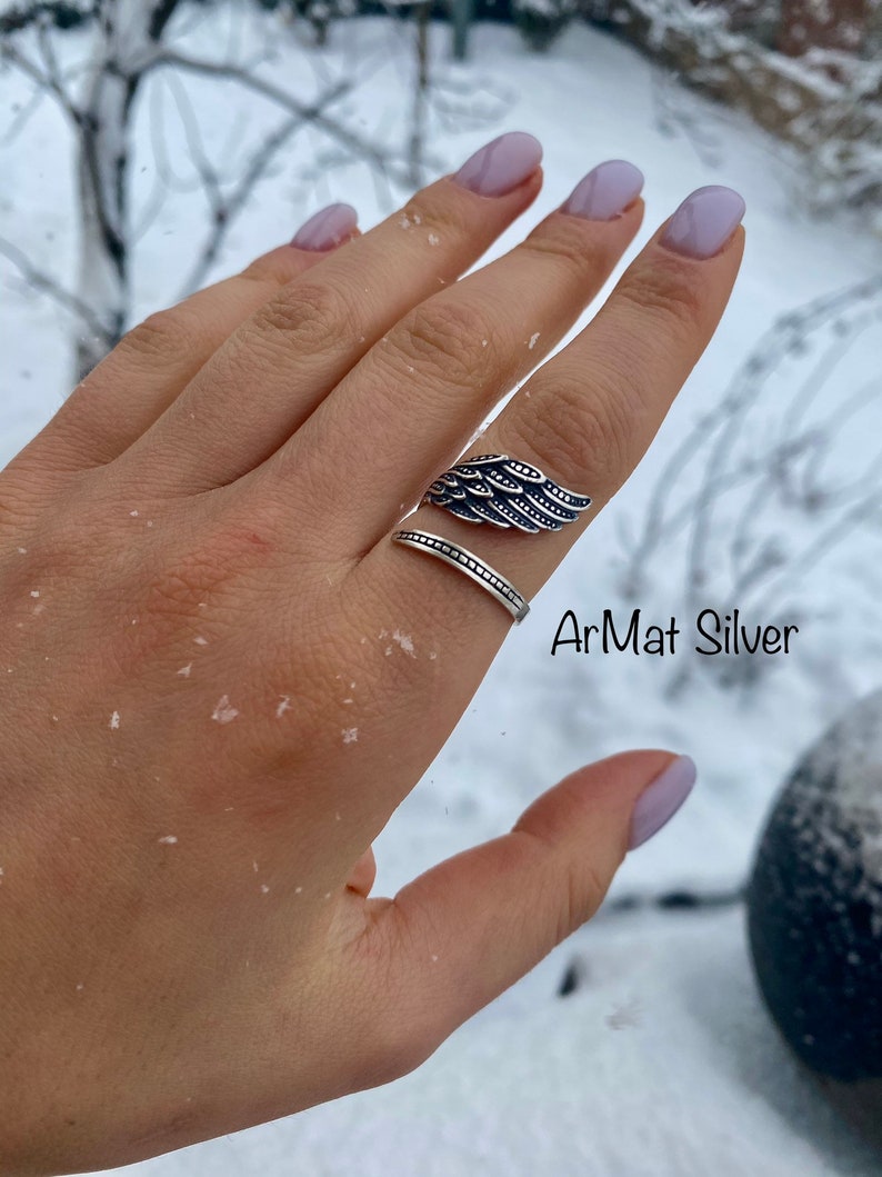 May include: A silver ring with a detailed wing design. The ring is worn on a finger with light pink nail polish. The text "ArMat Silver" is visible in the image.