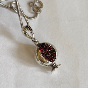 May include: A silver chain necklace with a pomegranate pendant. The pendant is made of silver and has red stones that resemble pomegranate seeds.